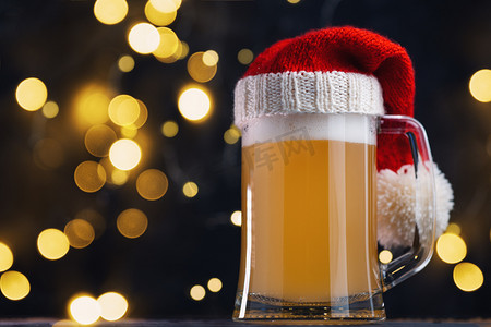 美国新年摄影照片_圣诞啤酒啤酒杯在圣诞老人的帽子黑暗的背景与bokeh花环。手工艺啤酒
