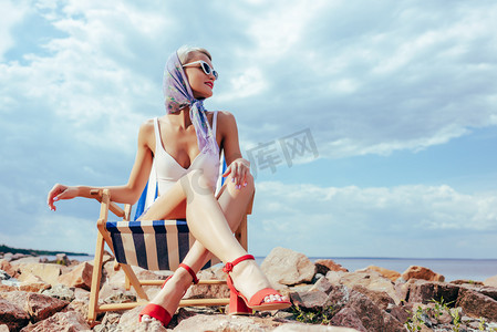 沙滩椅摄影照片_优雅的女人在复古泳装坐在沙滩椅在岩石岸边