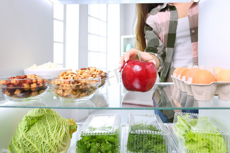 女性站在开着的冰箱旁边的画像，冰箱里装满了健康的食物、蔬菜和水果。女性的肖像