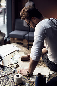 英俊的裁缝男性留着胡子，穿着白衬衣，与附近的木桌和线程和剪刀在古董家具与模特的惊人工作室工作背景的棕色皮制背带眼镜