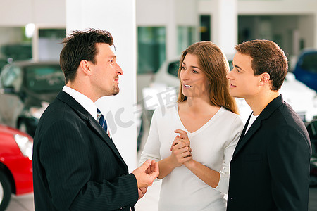 在一家汽车经销商的销售情况下，经销商正在与一对年轻夫妇交谈，背景中站着几辆车