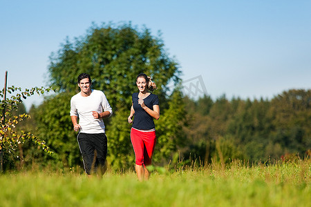 年轻的健身夫妇在户外做运动，在晴朗的蓝天下，在青草丛中的夏季草地上慢跑