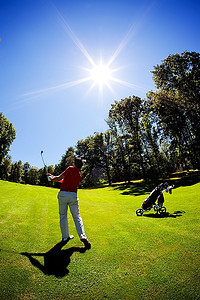 一名年轻的白人男子高尔夫球手，穿着红色衬衫和白色裤子，在球道上走向果岭。竖框，夏日晴朗，背光。