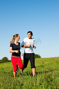 年轻的健身夫妇在户外做运动，在晴朗的蓝天下，在青草丛中的夏季草地上慢跑