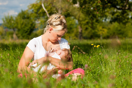 母亲母乳喂养她的宝宝在一个伟大的晴天在草地上有很多绿草和野花