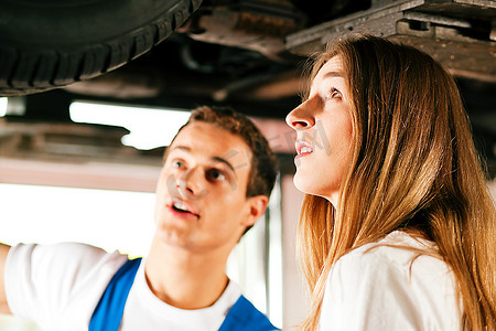 吊机摄影照片_一名妇女在他的修理厂与一名汽车修理工交谈，两人都站在汽车下面，这辆汽车是用汽车提升机吊起来的