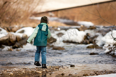 河边散步摄影照片_冬天在河边散步的小男孩；这个孩子在扔雪球