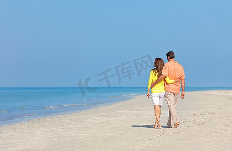身穿白色衣服的男女浪漫情侣在晴朗蓝天的荒凉热带海滩上漫步的背影