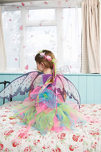年轻女孩(5-6岁)穿着仙女服装坐在靠窗后景的床上