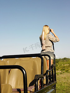 在狩猎中的年轻女子站在吉普车里，用双筒望远镜观察后景