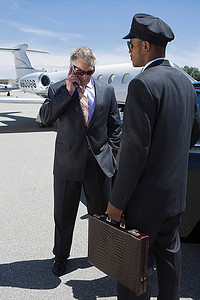 一位资深商人站在私人飞机外打电话，司机提着他的行李箱。