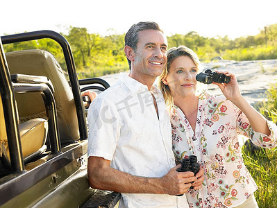一对夫妇站在吉普车旁，手持望远镜微笑着