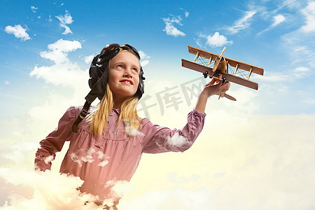 戴着飞行员头盔的小女孩在云的背景下玩玩具飞机