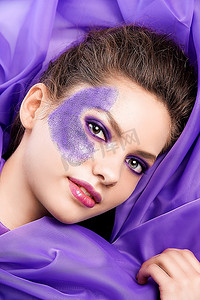 一位美女模特躺在紫色面料之间展示她闪闪发光的妆容的面部照片