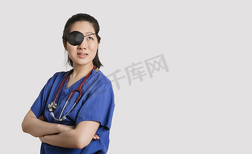 一名亚裔女医生戴着眼罩，双臂交叉在灰色背景上抬头看
