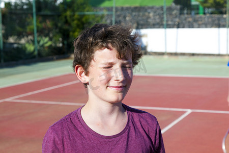 眼睛度假摄影照片_智能微笑的男孩在室外网球场，闭上眼睛