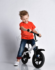 Young kid boy in orange t-shirt and blue jeans biker sits on his bicycle and looks at it like check if it is ready for race