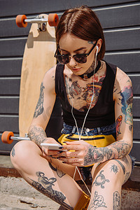 年轻纹身妇女在耳机听音乐与智能手机在滑板附近 