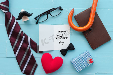 创意父亲节快乐摄影照片_快乐的父亲日概念。红领带, 眼镜, 胡子, 笔记本, 礼品盒与快乐的父亲节文本和手工红色的心在明亮的蓝色粉彩木桌背景.