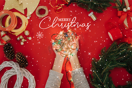 裁剪拍摄的妇女拿着圣诞树的灯在手与松树树枝和装饰手工花圈在红色的背景下, 用 