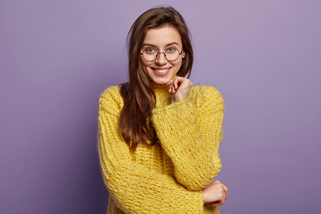 Optimistic pleasant looking woman with appealing appearance, keeps hands partly crossed over waist, listens intriguing interesting story, wears optical glasses and winter jumper, stands indoor
