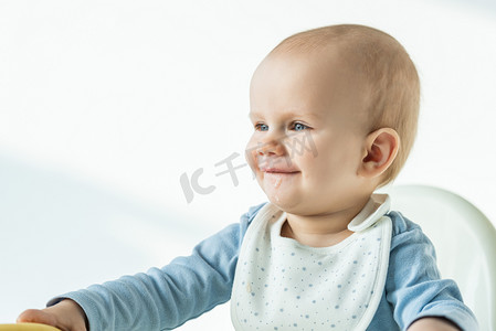 儿童baby摄影照片_Smiling baby boy with soiled mouth sitting on feeding chair on white background