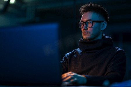 网络犯罪，黑客和科技概念-戴眼镜的年轻人在黑暗的房间里用笔记本电脑写代码。戴眼镜的年轻人在黑暗的房间里拿着笔记本电脑