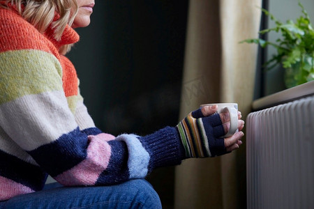 戴手套的女人与热饮料试图保持温暖的散热器在成本的生活能源危机