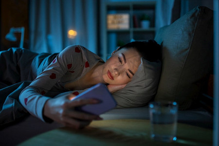 躺在床上摄影照片_技术，就寝时间和休息概念—十几岁的女孩在睡衣与智能手机躺在床上晚上。十几岁的女孩有电话躺在床上晚上