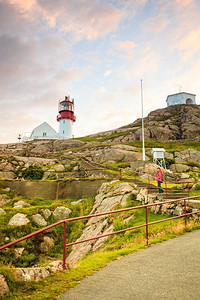 旅游女子和历史悠久的红白灯塔，位于挪威南部多岩石海岸的边缘，林德恩FYR灯塔。挪威的Lindesnes灯塔