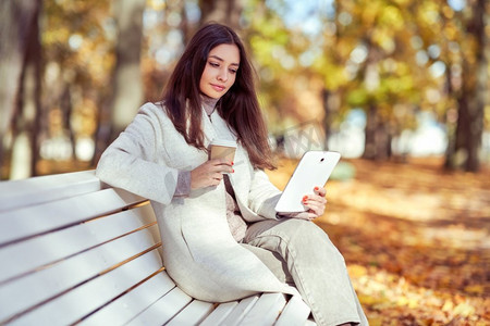 秋天公园里，一位年轻的美女坐在长椅上喝着咖啡。秋天的森林里，一个女孩在看电子书