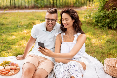 手机夏季夏季摄影照片_休闲和人的概念—快乐的夫妇与智能手机有野餐在夏季公园。愉快的夫妇与智能手机在野餐在公园