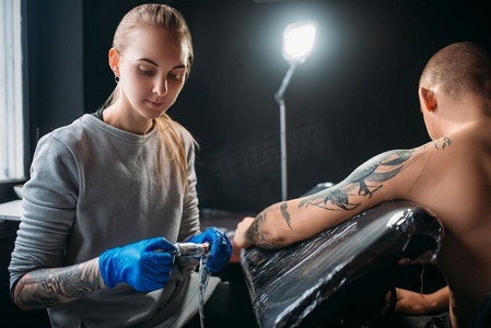 女纹身师用机器在肩膀上做纹身。专业纹身在沙龙。纹身师通过机器在男性肩膀上纹身