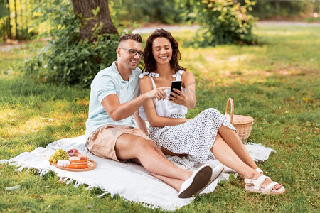 休闲和人的概念—快乐的夫妇与智能手机有野餐在夏季公园。愉快的夫妇与智能手机在野餐在公园