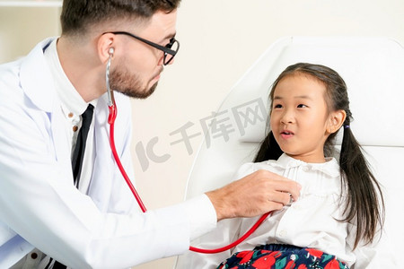 孩子住院摄影照片_年轻的男医生在医院办公室检查小孩。孩子很高兴，不怕医生。儿童医疗保健概念。