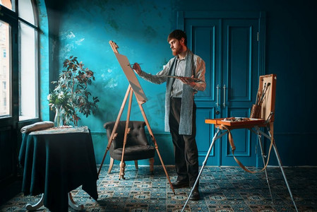 男性艺术家，在窗前的画架上用调色板和画笔手绘。油画、画笔画