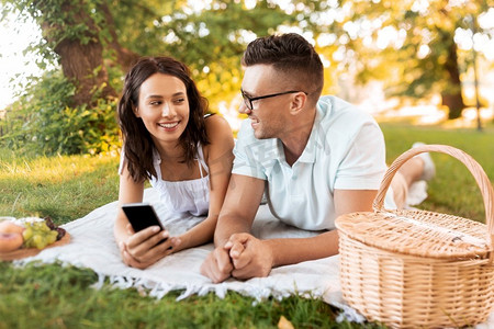 手机夏季夏季摄影照片_休闲与人的概念快乐夫妇带着智能手机在夏季公园野餐。带着智能手机在公园野餐的幸福情侣