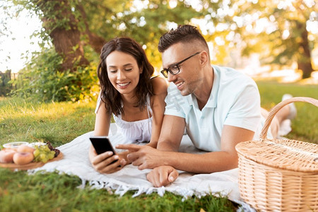 休闲和人的概念—快乐的夫妇与智能手机有野餐在夏季公园。愉快的夫妇与智能手机在野餐在公园