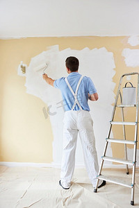 泼出去的油漆摄影照片_后视图穿着工作服绘画墙壁在房间的房子与油漆滚筒