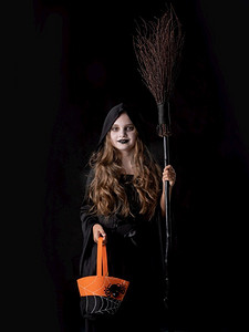 穿着万圣节女巫服装的女孩拿着扫帚和橙色袋子，在黑色背景上捉弄或治疗孤立的人。穿着万圣节服装的女孩不给糖就捣蛋