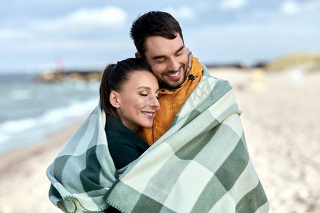 秋天爱情摄影照片_爱情，关系和人的概念-秋天海滩上，幸福的微笑着的情侣在温暖的毯子里。秋天海滩上裹着温暖毯子的幸福夫妇