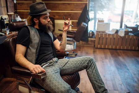 剪刀手icon摄影照片_残忍的胡子理发师在帽子与直剃刀手坐在皮椅子，理发店inior在背景