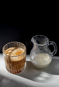 一杯牛奶杯摄影照片_咖啡因，物品和饮料概念-桌上有一杯咖啡和加牛奶或奶油的罐子。桌上有玻璃杯咖啡和一罐牛奶或奶油