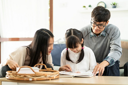 卡通带逗号字体摄影照片_亚洲可爱的女孩阅读故事卡通书与她的妈妈和爸爸在客厅。幸福快乐的家庭和家庭生活理念。