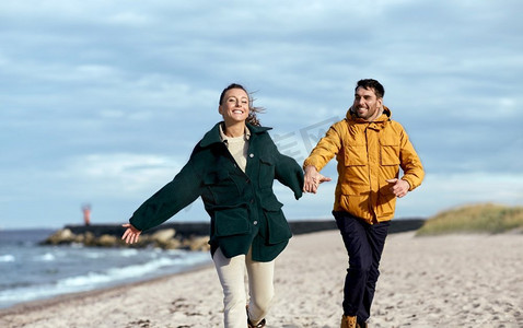 爱情、关系和人的概念-幸福的微笑情侣沿着秋日海滩奔跑，手牵手。一对情侣沿着秋天的海滩跑步