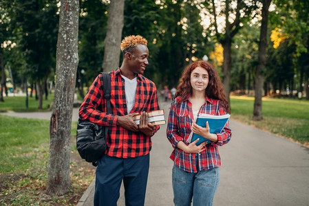 人走摄影照片_两个学生走在夏季公园的人行道上。男性和女性白人青少年在户外放松。两名学生走在夏季公园的人行道上