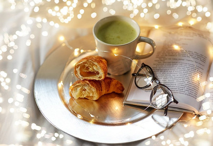 早上，hygge和早餐的概念—羊角面包，一杯抹茶茶书和眼镜在床上在家里。羊角面包，抹茶茶，书和眼镜在床上