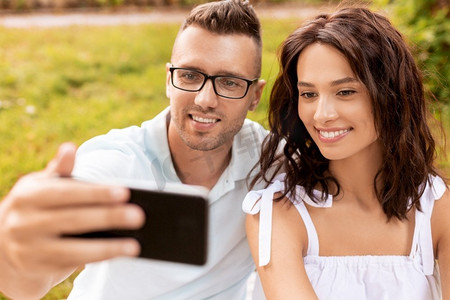 手机夏季夏季摄影照片_休闲和人的概念—快乐的夫妇野餐和采取自拍与智能手机在夏季公园。快乐的夫妇采取自拍在夏季公园