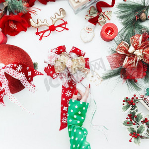 圣诞节装饰摄影照片_穿着绿色衬衫的女性手持圣诞蝴蝶结在白色桌面上，上面有红色的节日装饰。顶视图