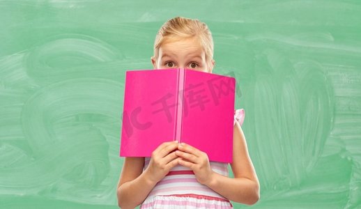 绿色阅读摄影照片_童年和人的概念—隐藏在书后面的小女孩在绿色黑板背景小女孩躲在书在学校